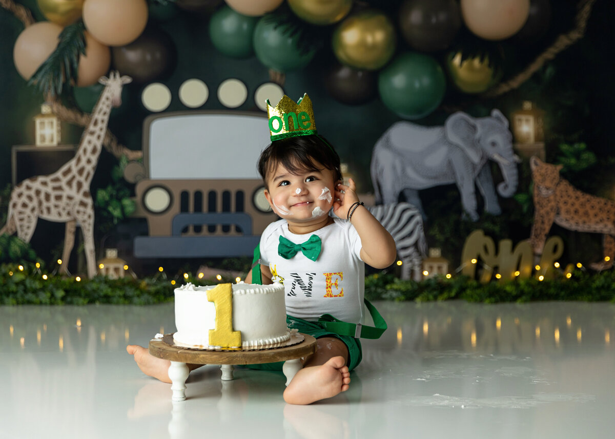 A toddler boy in green suspenders smiles in his jungle themed birthday photos