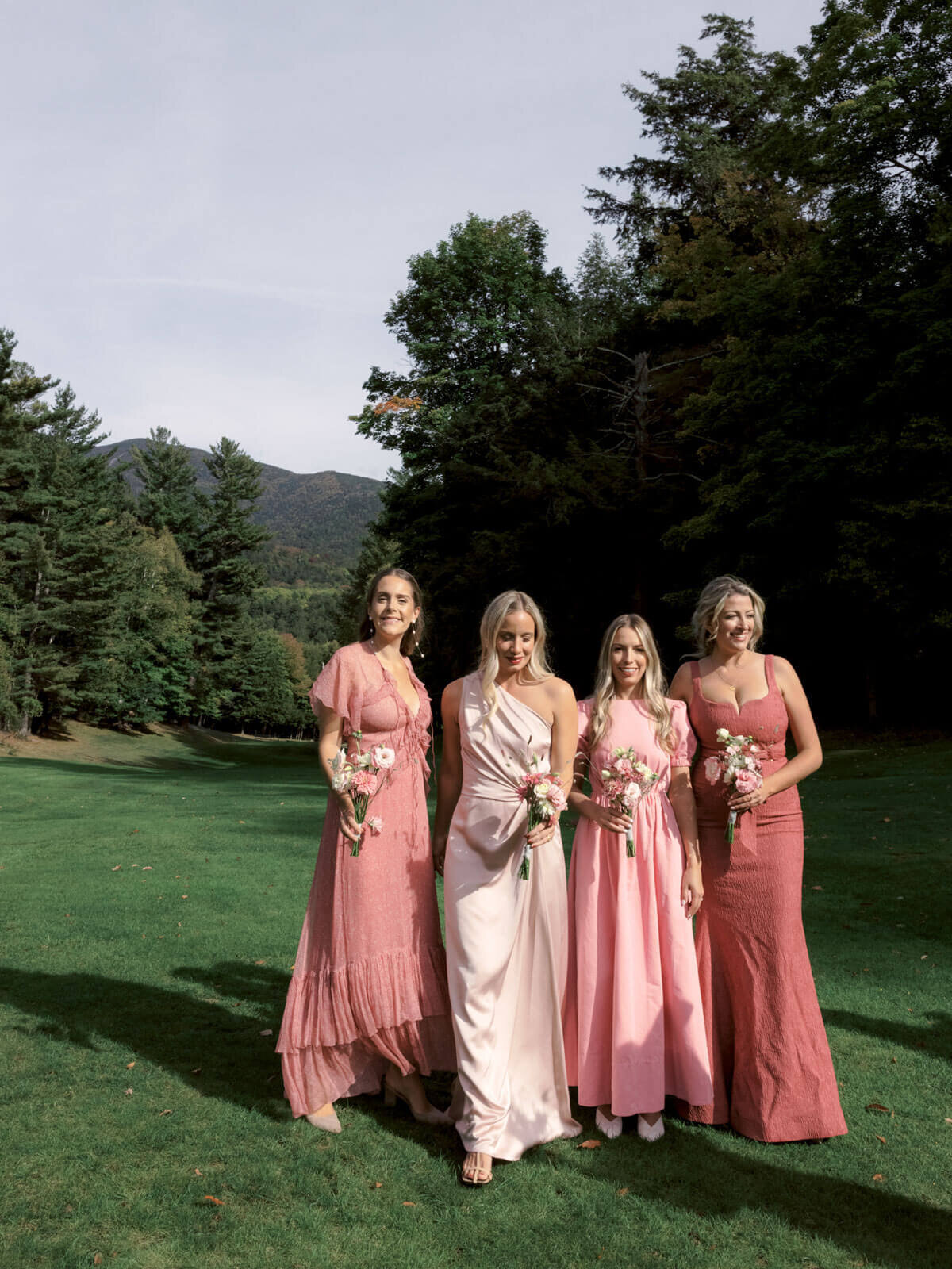 Four bridesmaids in different shades of peach, standing outdoors, trees behind, at The Ausable Club, NY. Image by Jenny Fu Studio.