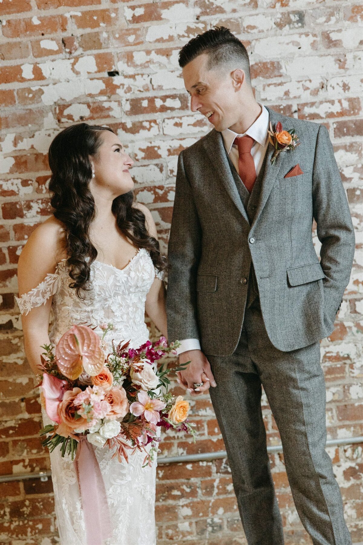 Bride wearing lace dress holding colorful bouquet and groom  wearing grey suit standing against brick wall  at the St Vrain, Longmont CO.