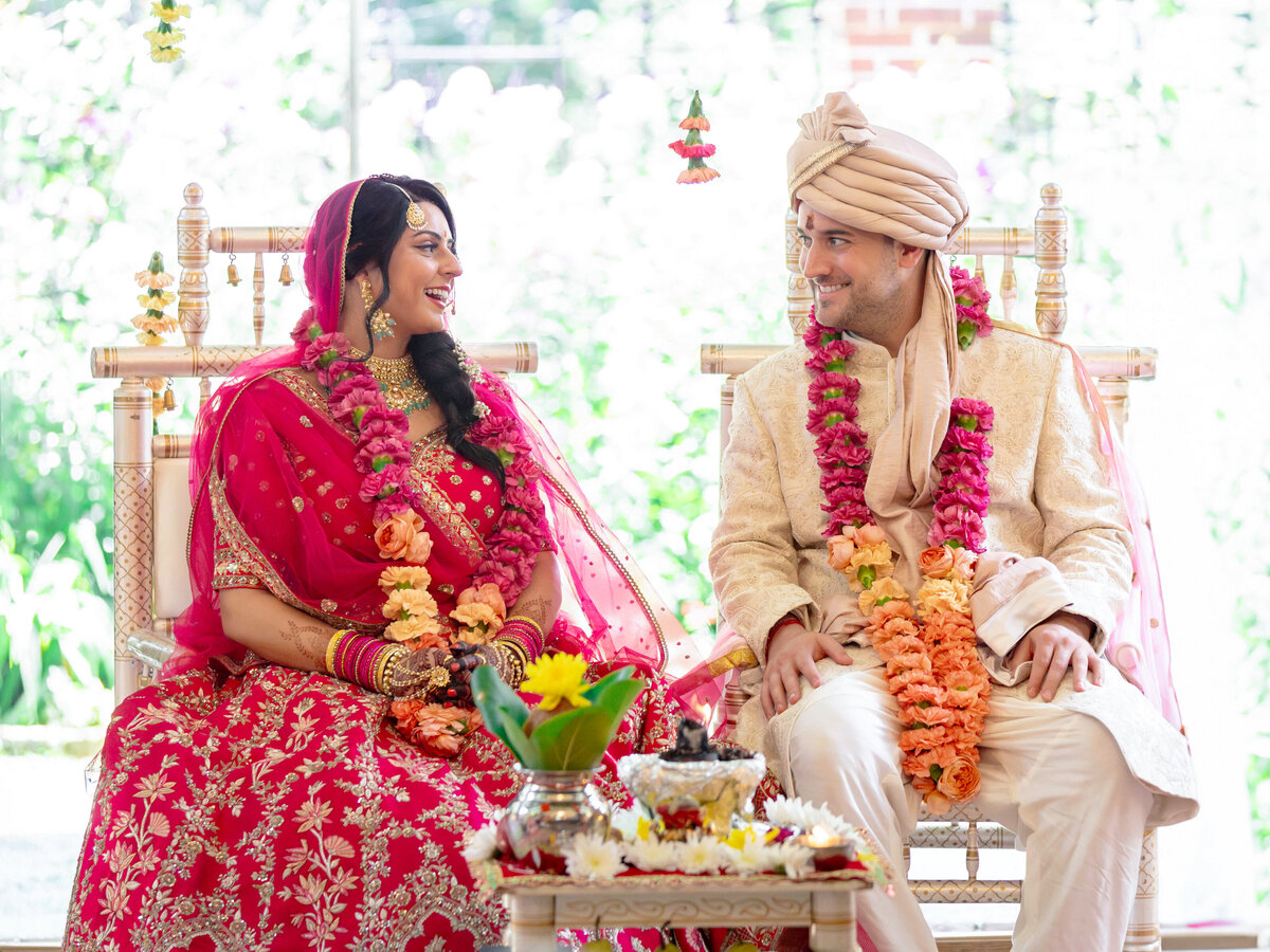 A bride and groom in traditional Indian wedding attire sit on ornate chairs, smiling at each other. Both are adorned with floral garlands. A small table with flowers and ceremonial items is in the foreground. The background is softly lit.