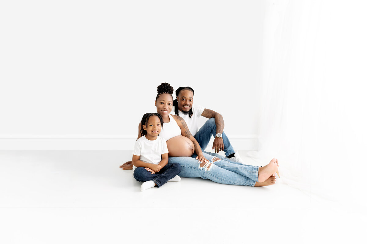 Happy expecting parents sit on the floor of a Atlanta Maternity Photographer studio with their toddler all in jeans and white shirts
