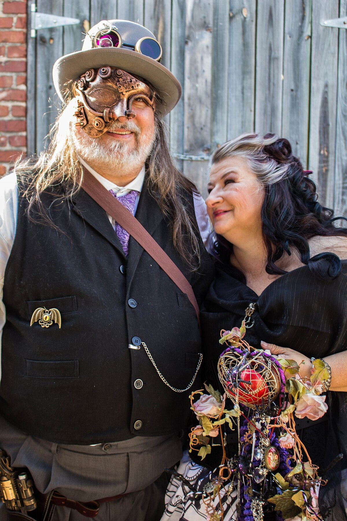 The steampunk groom wearing grey pants, a black vest with a white shirt and purple tie has a very detailed clockwork half mask and fedora hat with tinted steampunk goggles. The bride, wearing a black and white wedding dress to match her black and white hair holds a bouquet of broken watch pieces, brooches, skeleton keys and blush roses.