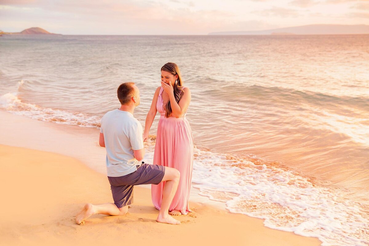 Woman in pink maternity gown holds her hand over her mouth in surprise during proposal session with Love + Water on the island of Maui