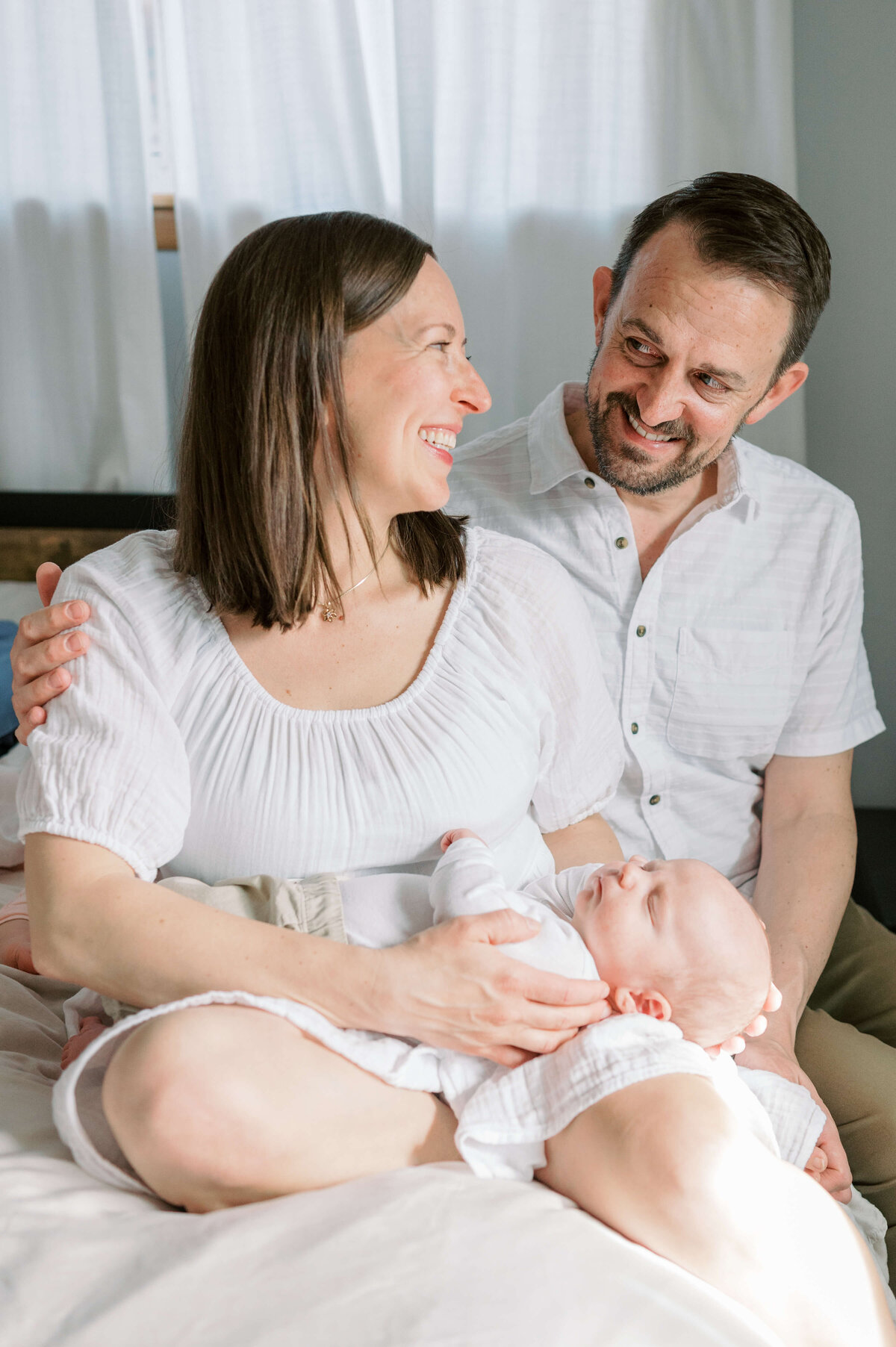 A smiling woman is hugged by her husband as she holds their new son in her lap in an image by Virginia family photographer