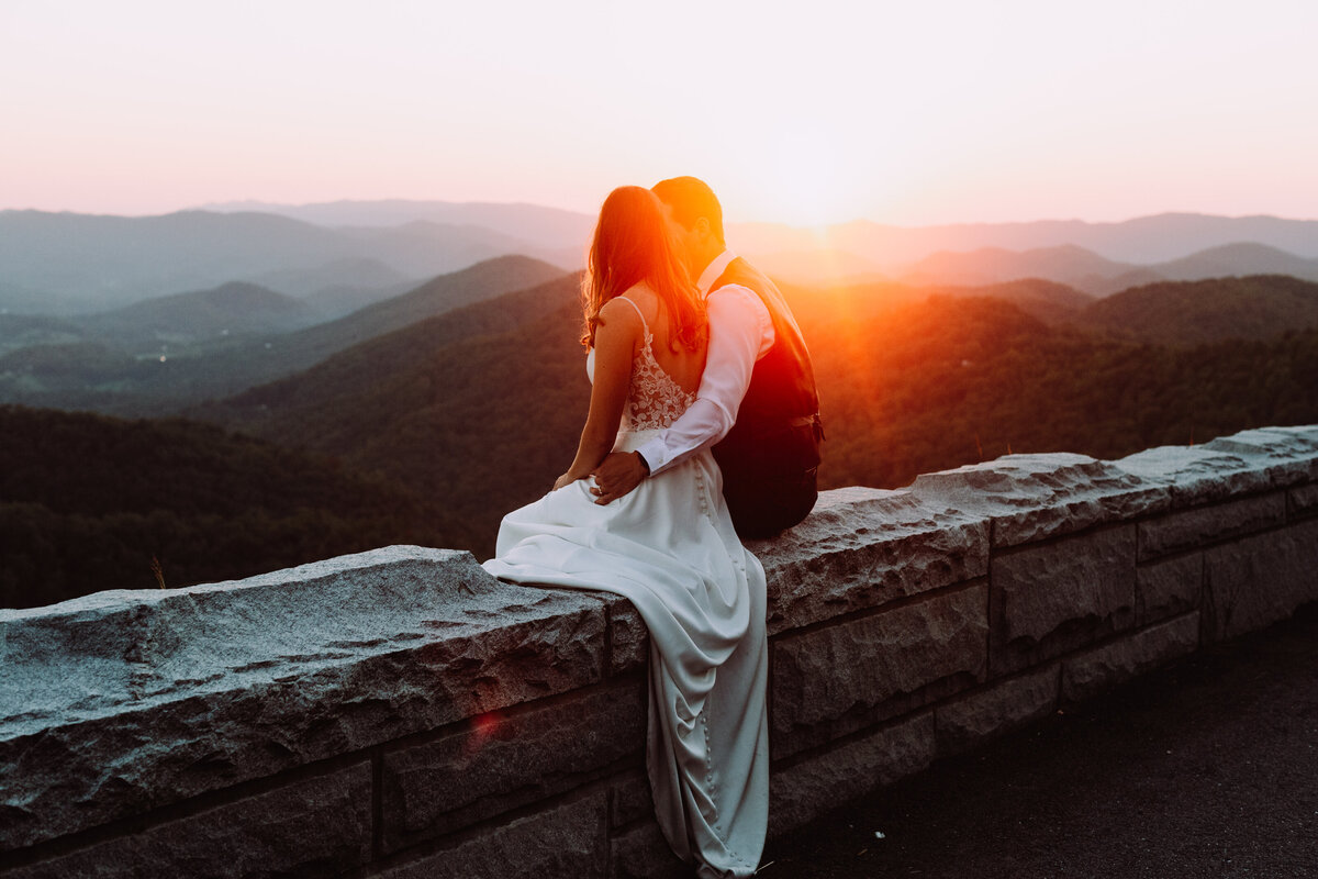 Bride and Groom sitting on a wall while the sun setting over the mountains. Your Love Deserves a Scenic Backdrop