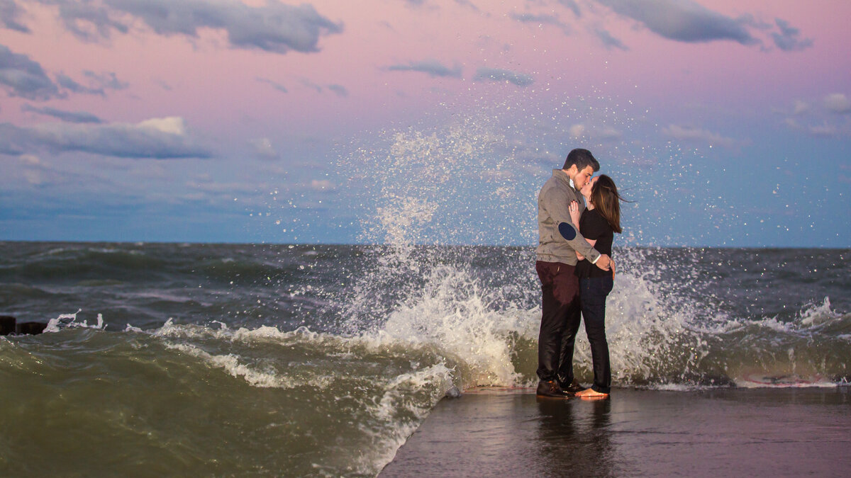 Waves crash over a couple taking sunset engagement photos in Chicago.