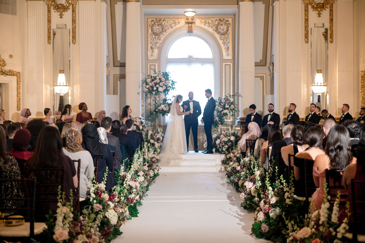 A bride and groom stand facing each other at the altar in an ornate hall. The aisle is lined with floral arrangements, and guests are seated on either side. A large window and decorative pillars create an elegant backdrop.