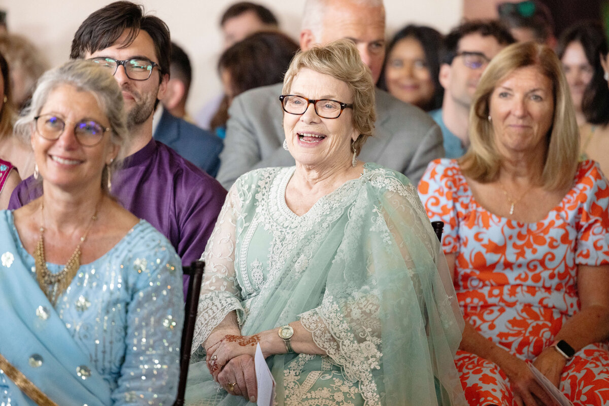 A group of people sitting and smiling at an event. An older woman in the center wears a light blue and silver outfit. To her left, another woman in glasses and a patterned blue shirt smiles. The audience is engaged and cheerful.