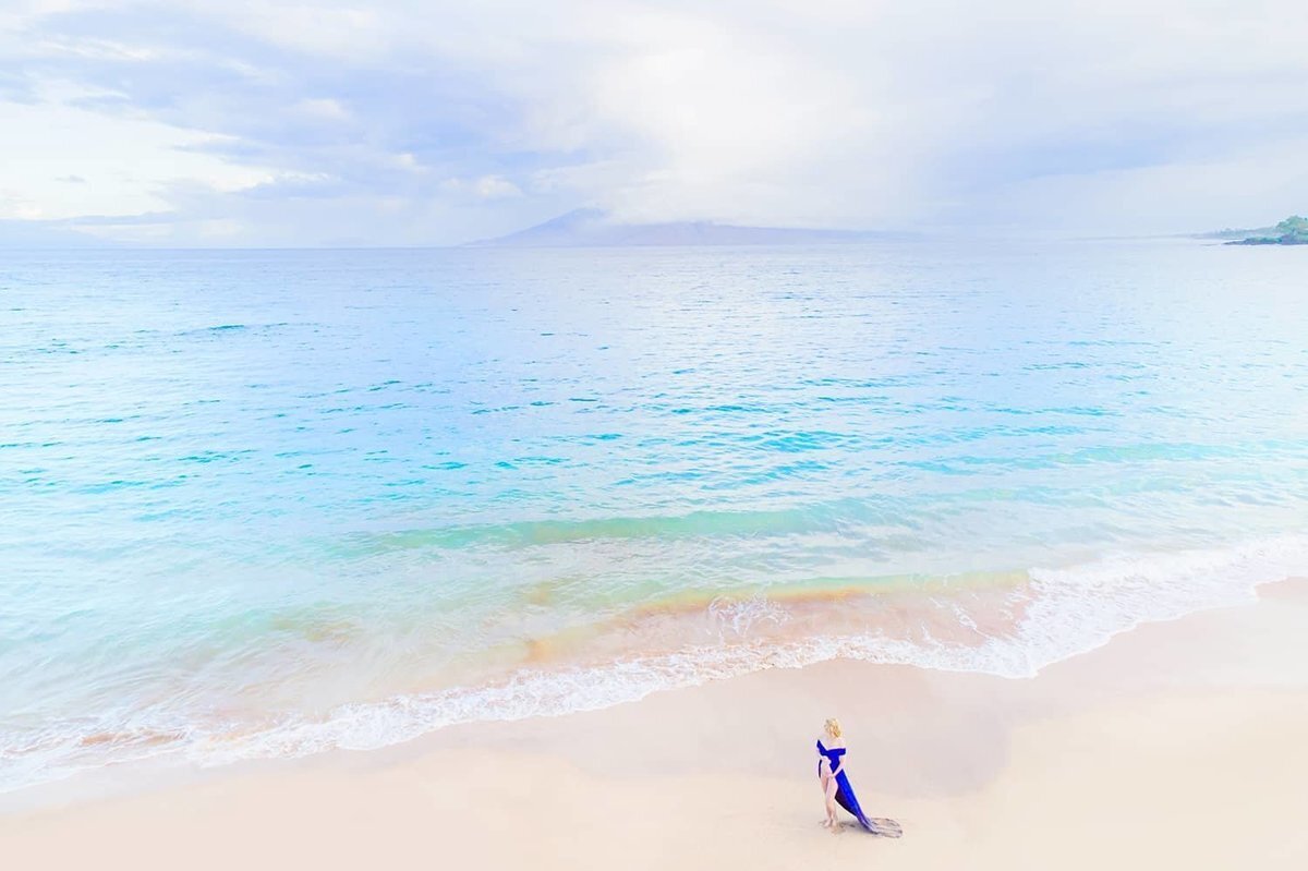Drone portrait of pregnant woman wearing a jewel toned maternity gown. She stares out to sea as the drone flies overhead.