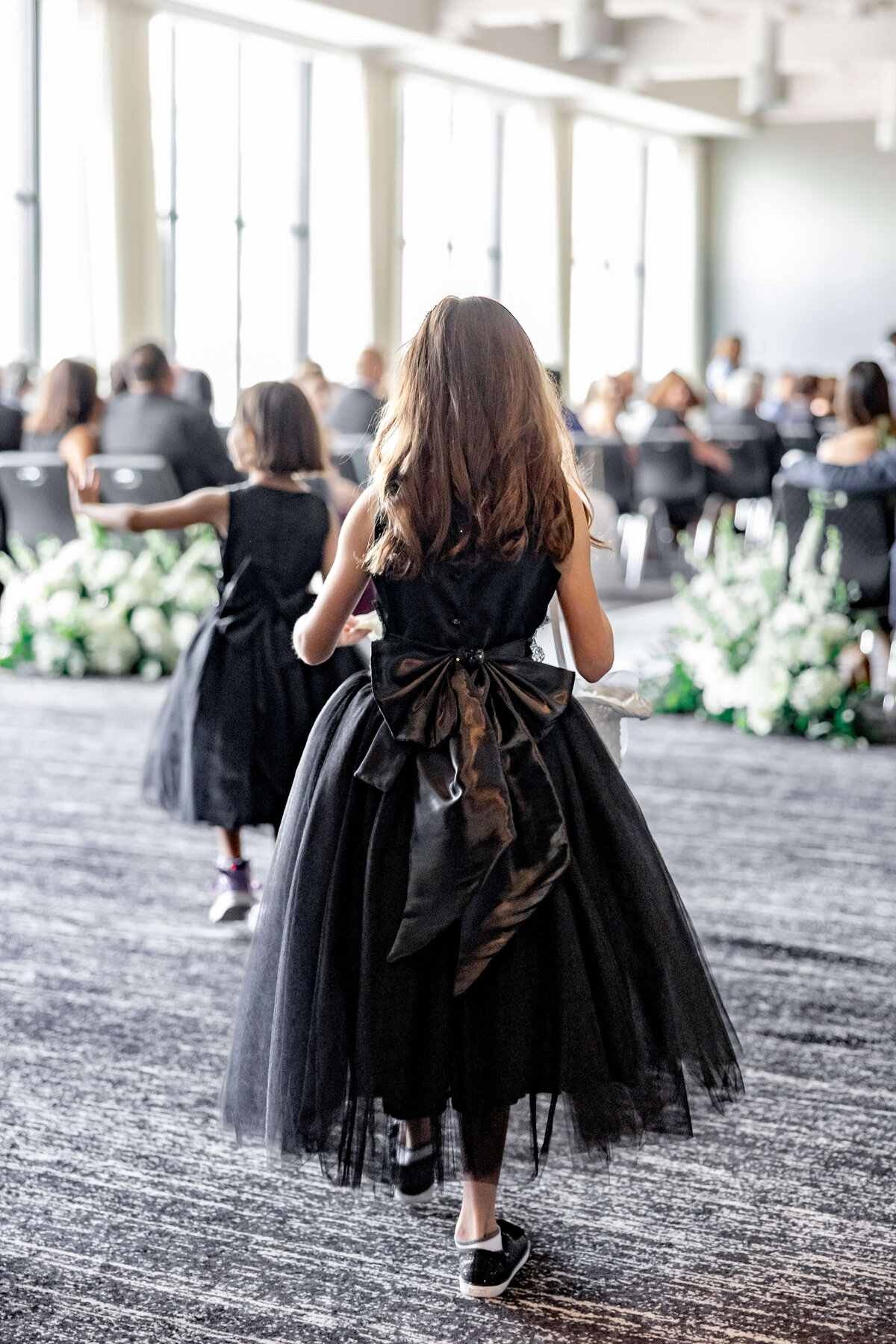 Two young girls in black dresses with bows walk down an aisle at a formal event. The setting has large windows and floral arrangements, and many seated guests are visible in the background.