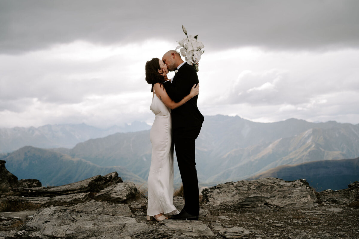 The Lovers Elopement Co - wedding couple kiss on top of mountain, with mountains in background