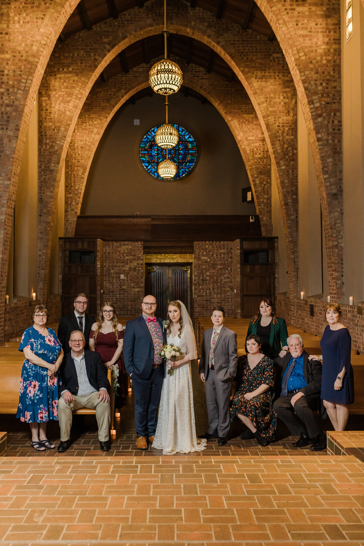 Portrait of a bride, groom, and their families after their wedding ceremony at the Little Chapel in the Woods at Texas Woman's University in Denton, Texas. The bride is on the right and is wearing an intricate white dress with a long veil while holding a bouquet. The groom is on the left and is wearing a navy suit with a patterned dress shirt and bowtie. Both families flank the couple on either side all in wedding guest attire. They whole group is backed by the intricate and intimate architecture of the chapel.