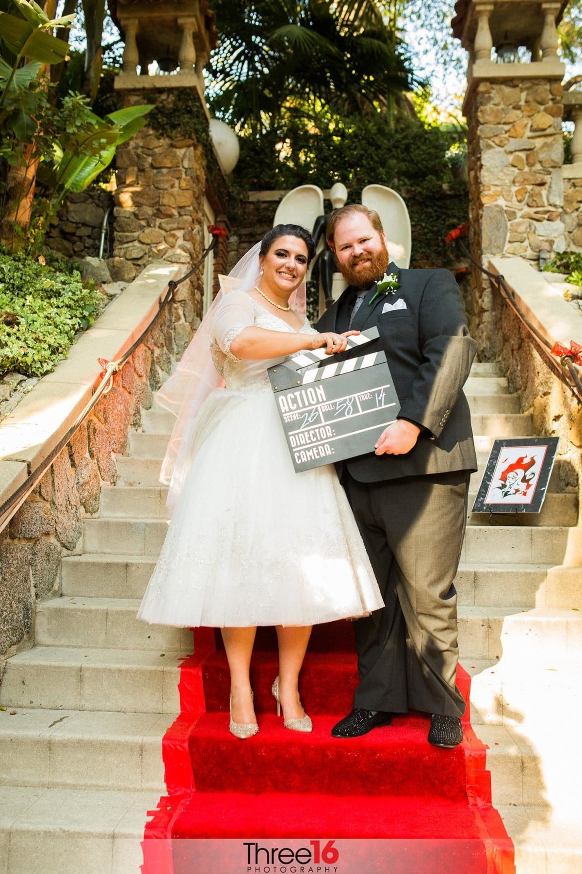 Bride and Groom pose on a a red carpet on stairs with a movie director sign
