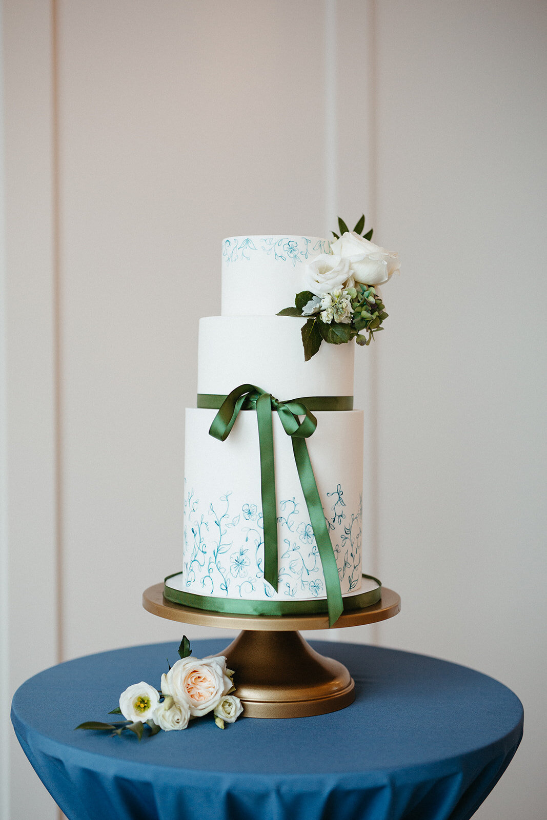 White cake with green ribbon and flowers on a gold cake stand atop a rounded table with blue linen.