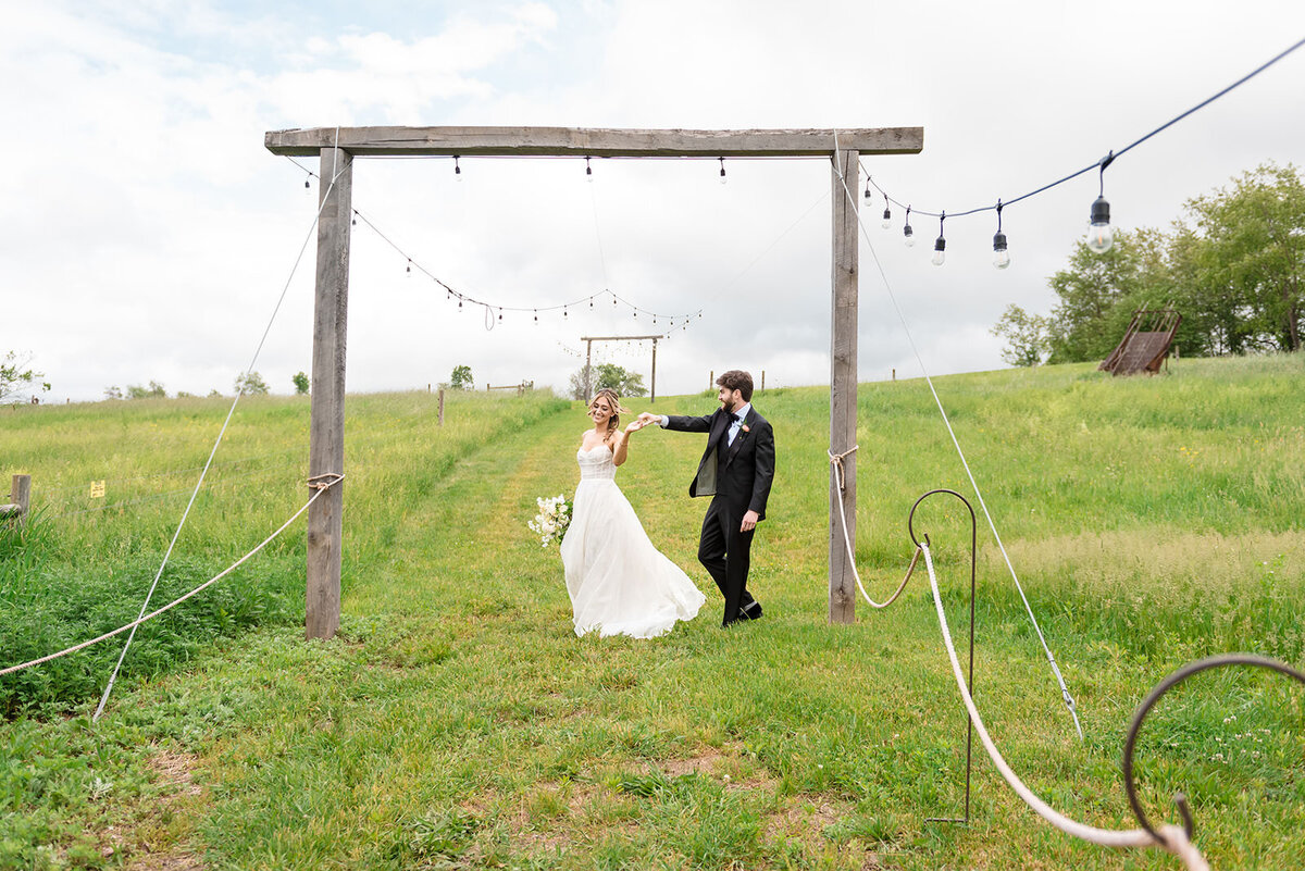 A bride leading the groom by the hand under a wooden structure adorned with string lights in a grassy field.