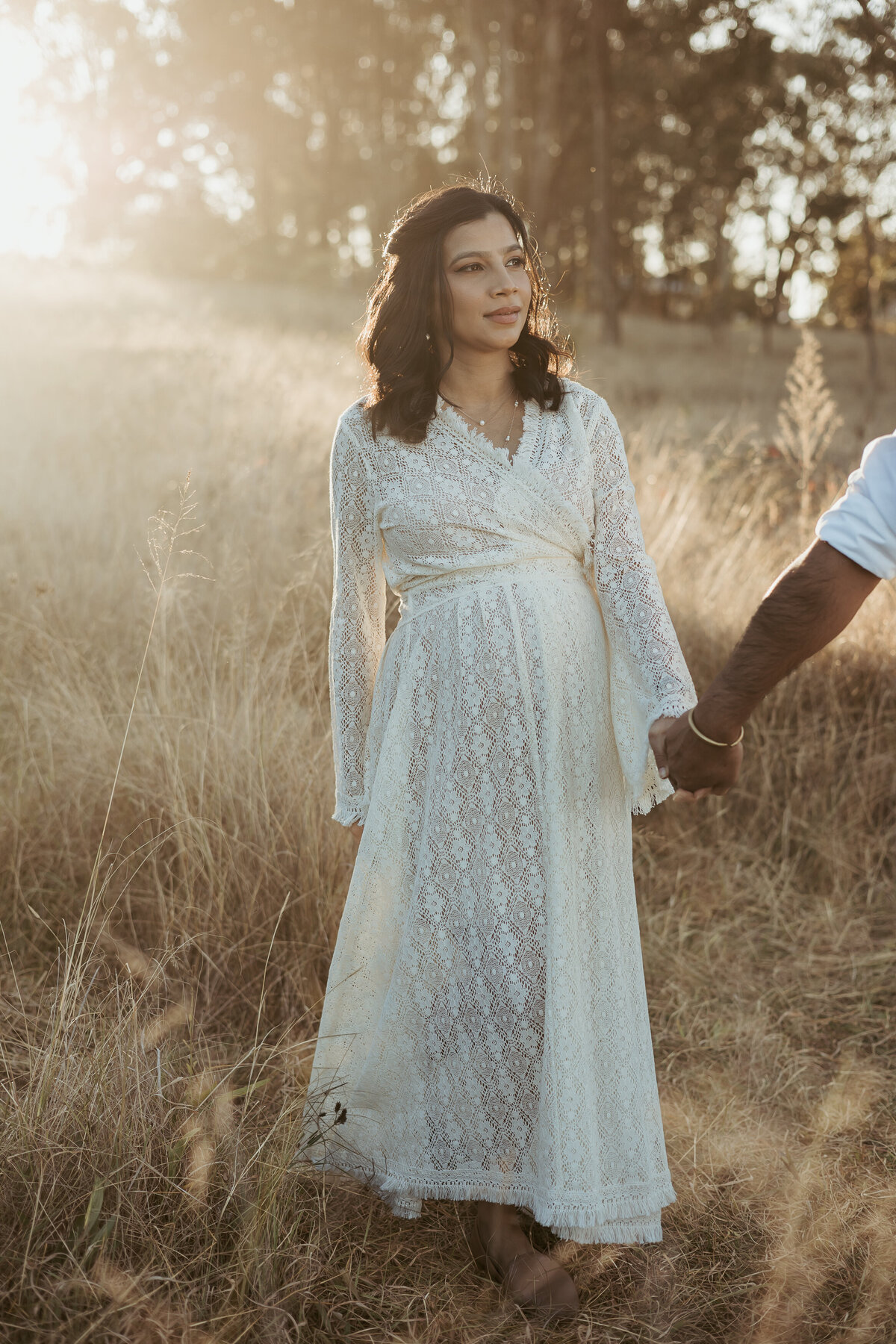 Mama wearing a white lace gown being led through a gorgeous field at sunset by her husband for their maternity photoshoot