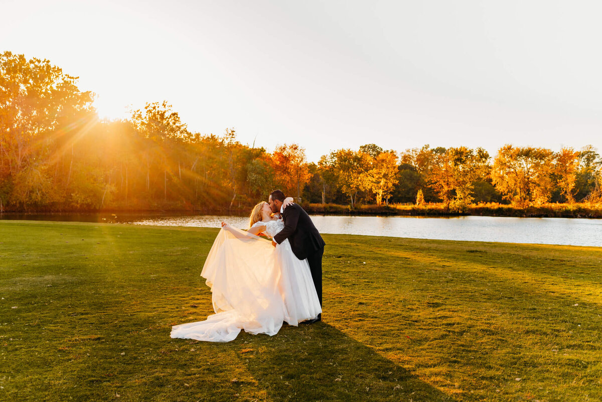 photo of a bride throwing the train of her dress as the groom kisses her in front of a pond at sunset