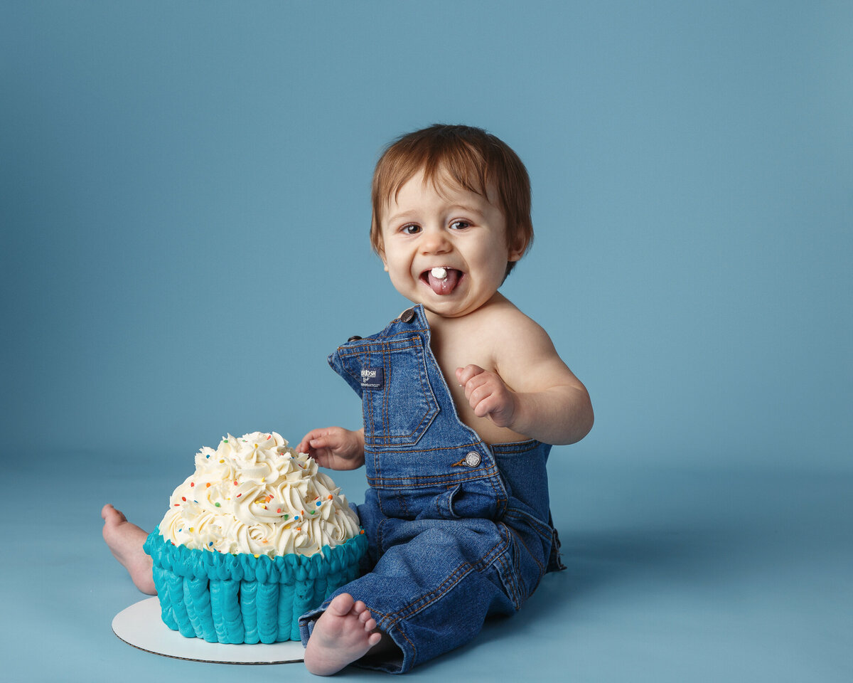 Milestone portrait of a one year old wearing overalls and sitting with a giant cupcake photographed by baby photographer