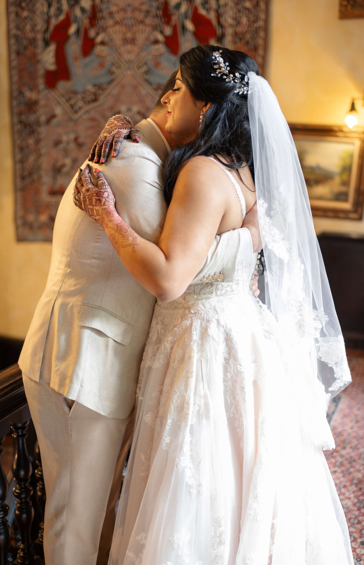 A bride in a white dress with a veil embraces a groom in a light-colored suit. The scene is set indoors with a patterned carpet. The brides hands have henna designs, and there is a tapestry hanging on the wall behind them.