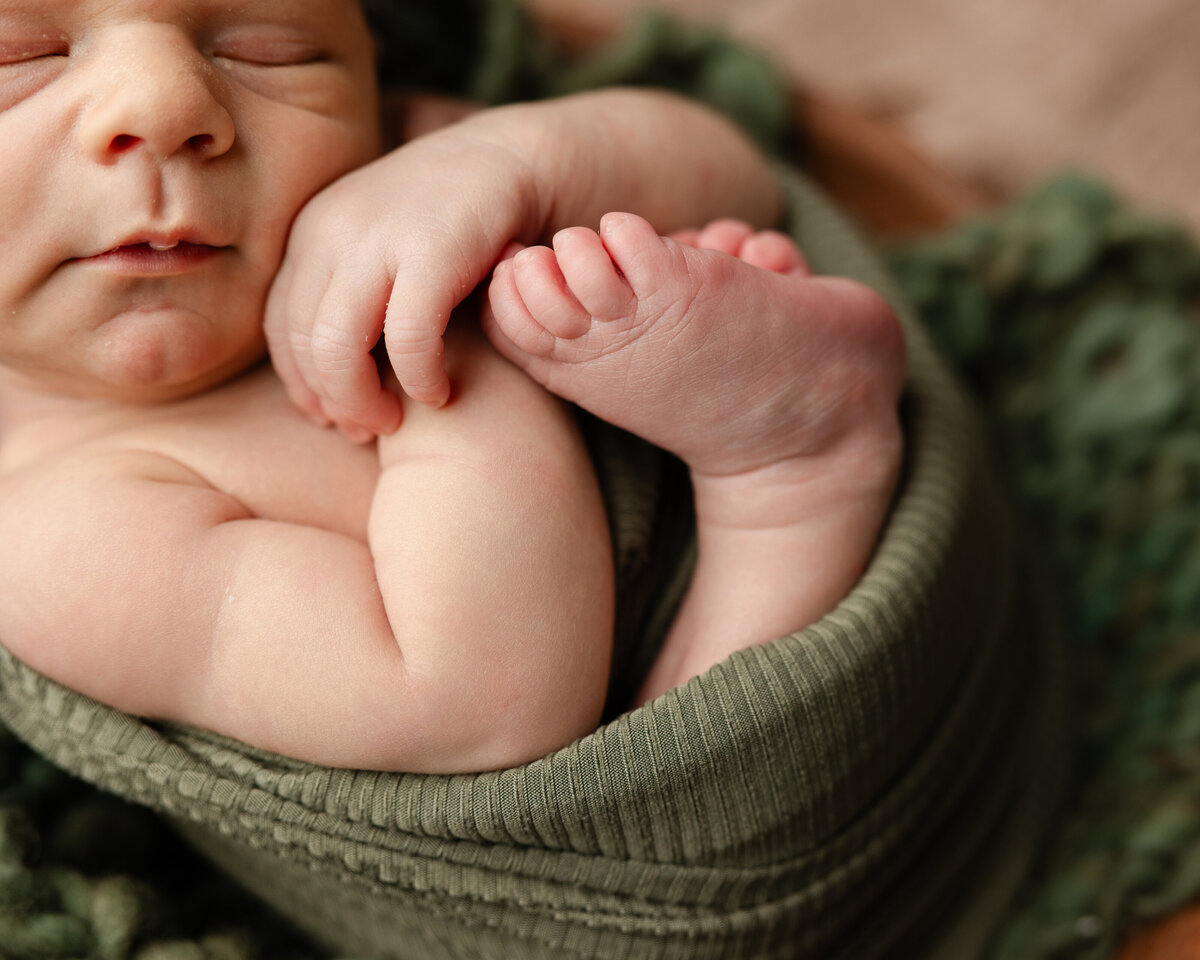 Close-up of a newborn baby boy’s curled hands and feet, wrapped in deep green fabric during a rustic bowl pose. Captured by Inland Empire newborn photographer Kaitlyn Dawn Photography.