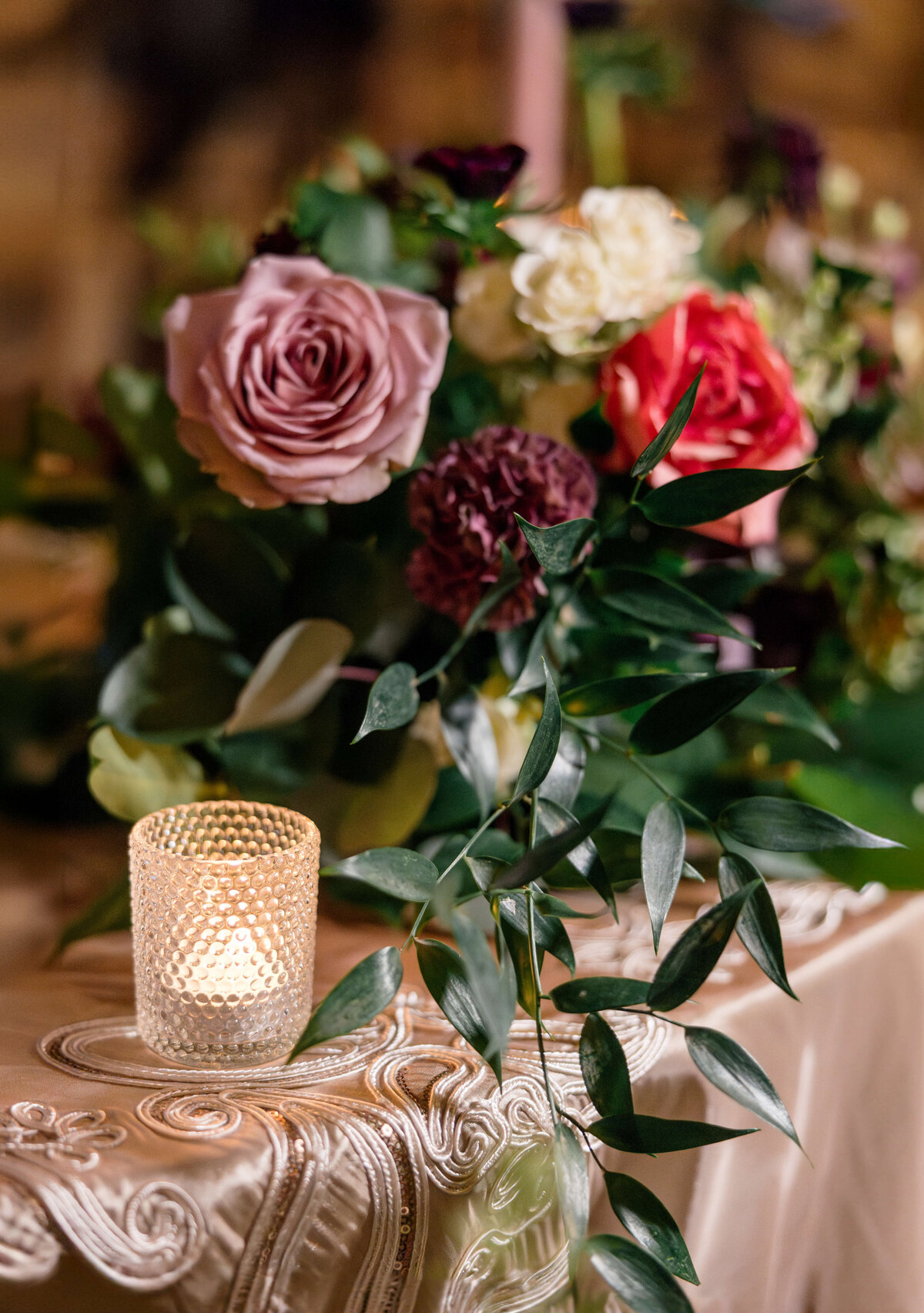 A softly lit candle in a textured glass holder sits on an ornate tablecloth, accompanied by an arrangement of pink, red, and white roses with dark foliage, creating an elegant and romantic atmosphere.