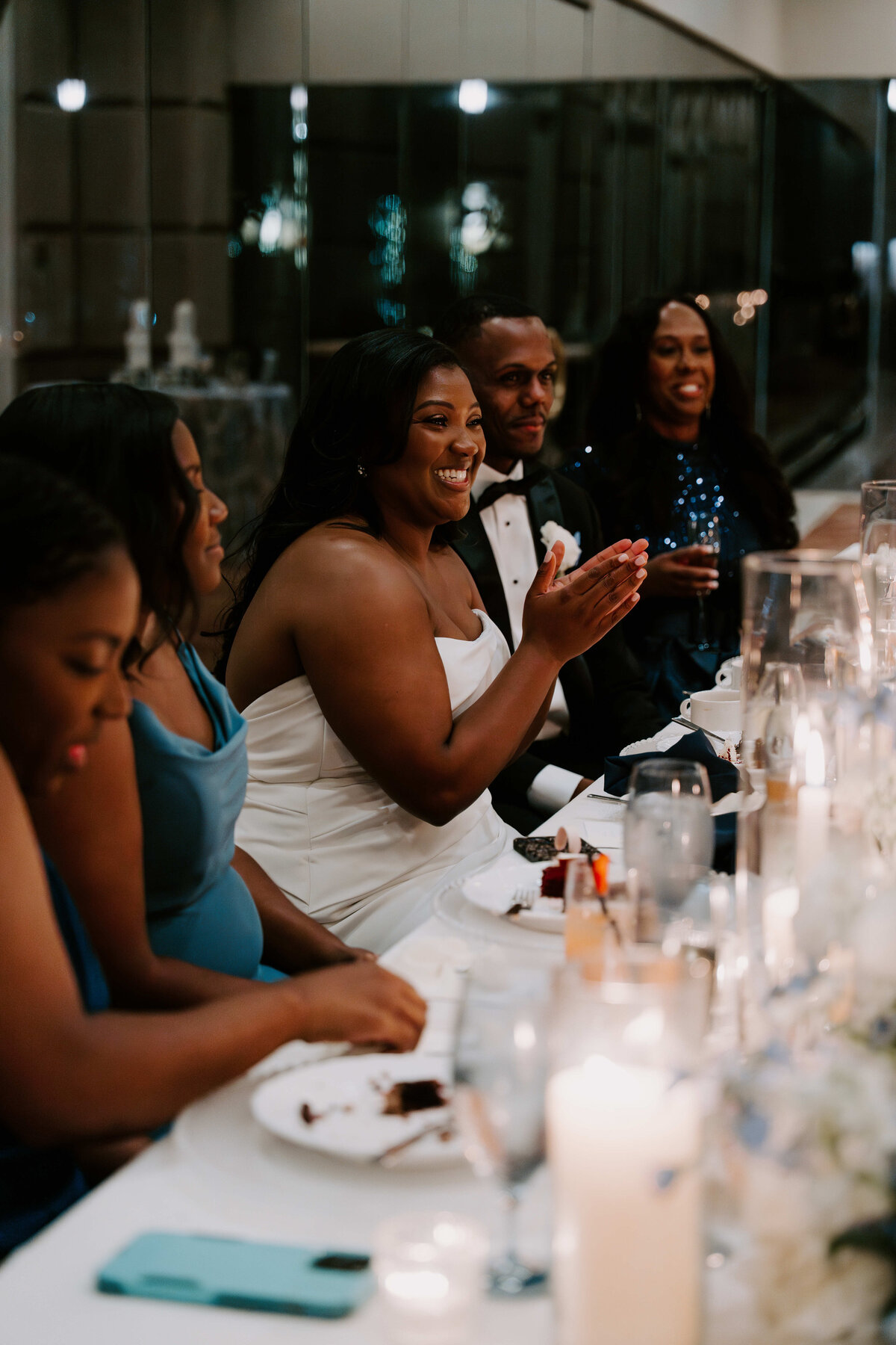 A joyful bride in a white dress claps while seated at a candlelit wedding reception table. Guests beside her in formal attire, creating a warm, celebratory atmosphere.