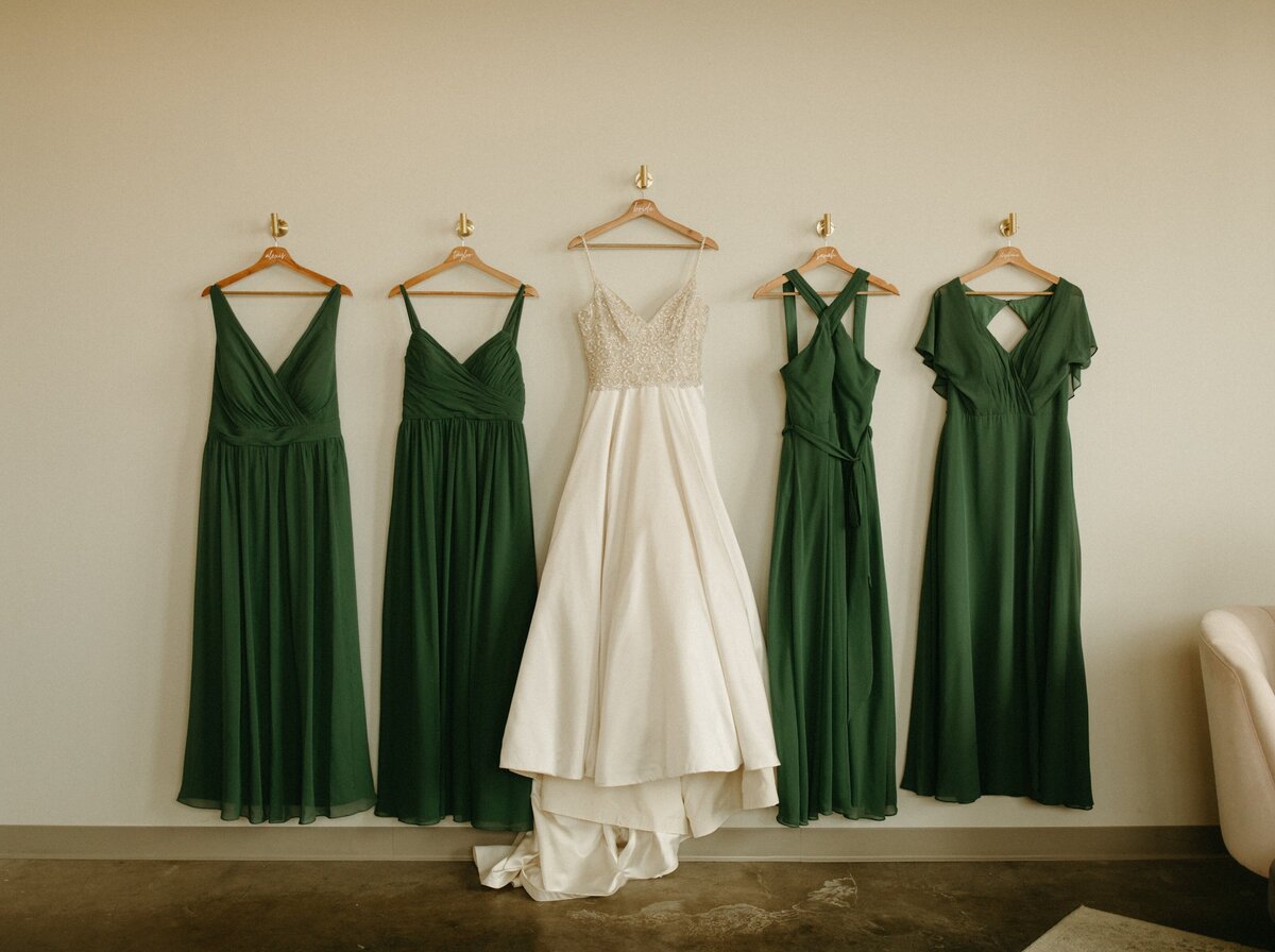 Four dresses hanging on a wall, with a white wedding dress in the center flanked by three green bridesmaid dresses, perfect for park farm winery weddings.