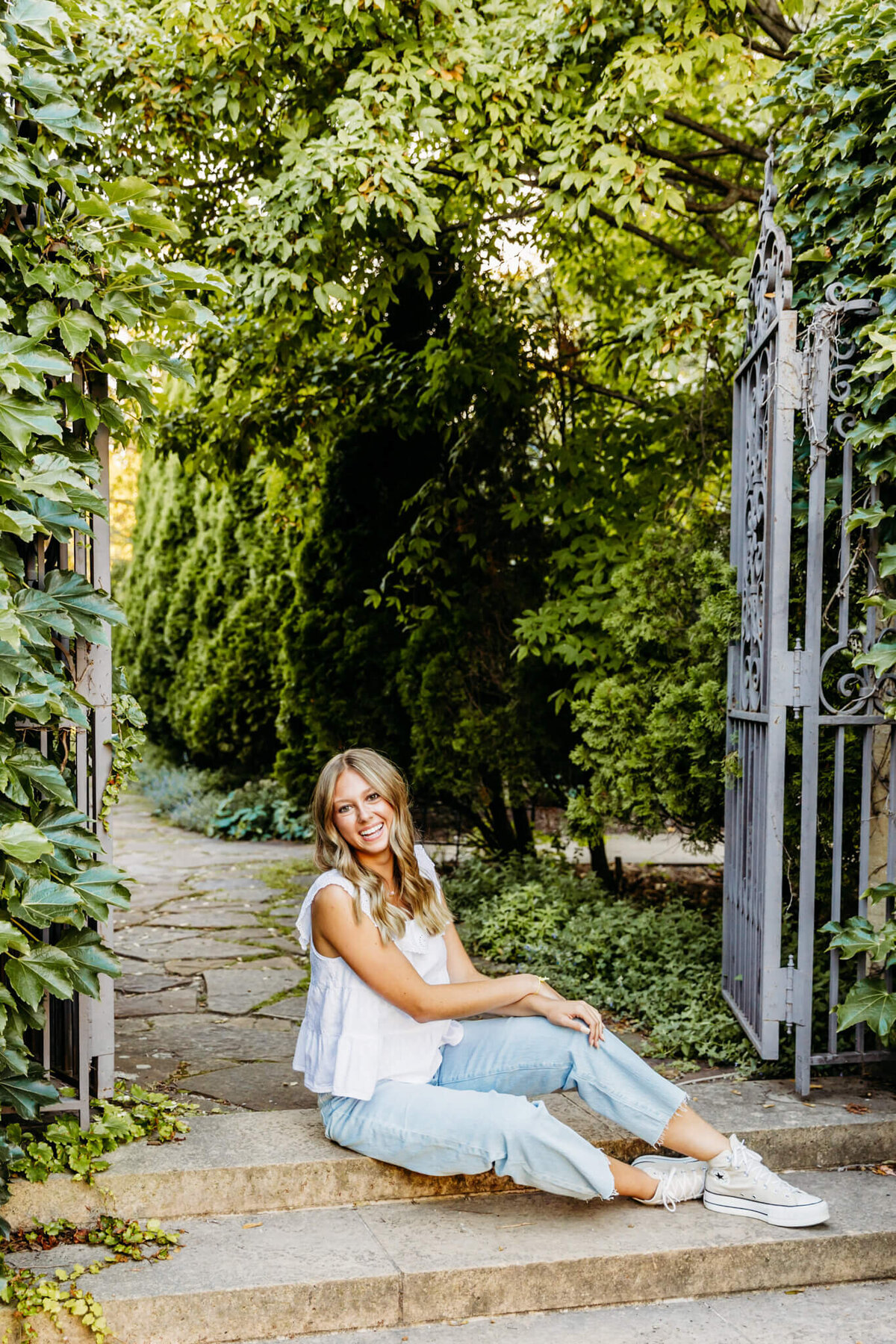 female high schooler in a white top sitting with her legs stretched out on stone steps in front of a black garden gate captured by Ashley Kalbus Photography