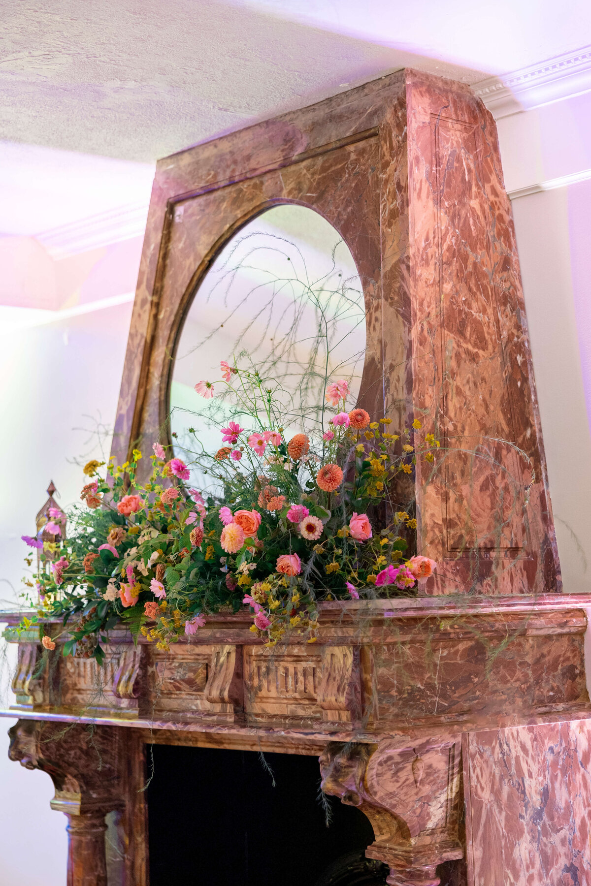 A tall, ornate marble fireplace with a large mirror above it, adorned with an arrangement of colorful flowers and greenery. The room is softly lit, highlighting the intricate details of the marble and the vibrant floral display.