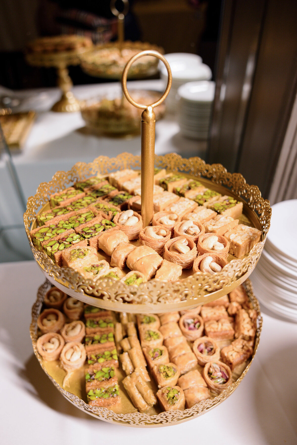 A two-tiered gold tray displays an assortment of pastries and desserts, including layered and rolled confections topped with nuts. The tray is adorned with an ornate edge and stands on a white table, with additional plates in the background.