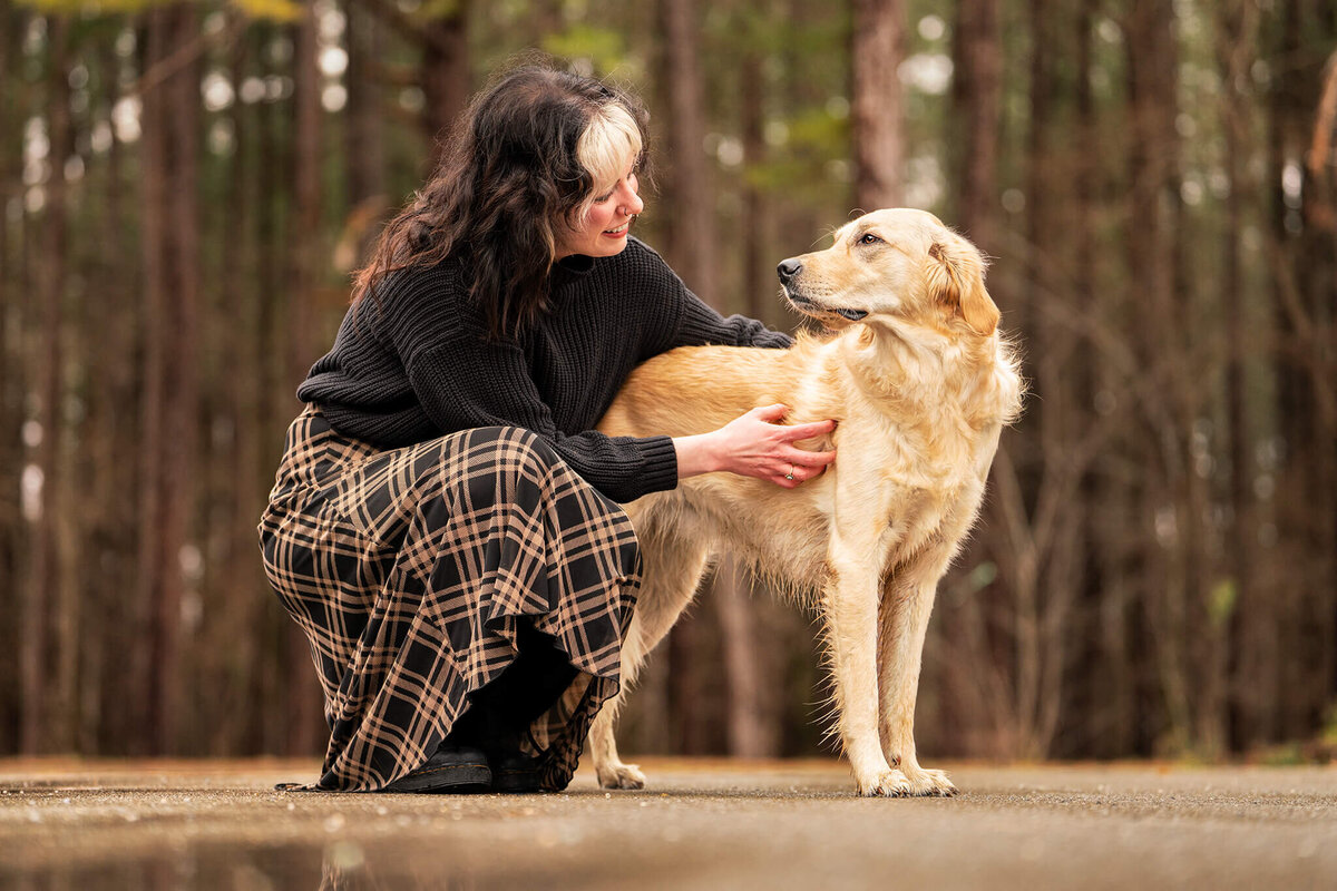 Dog owner in long plaid shirt with black tights and black boots crouching next to Golden Retriever dog sharing a loving look and embrace