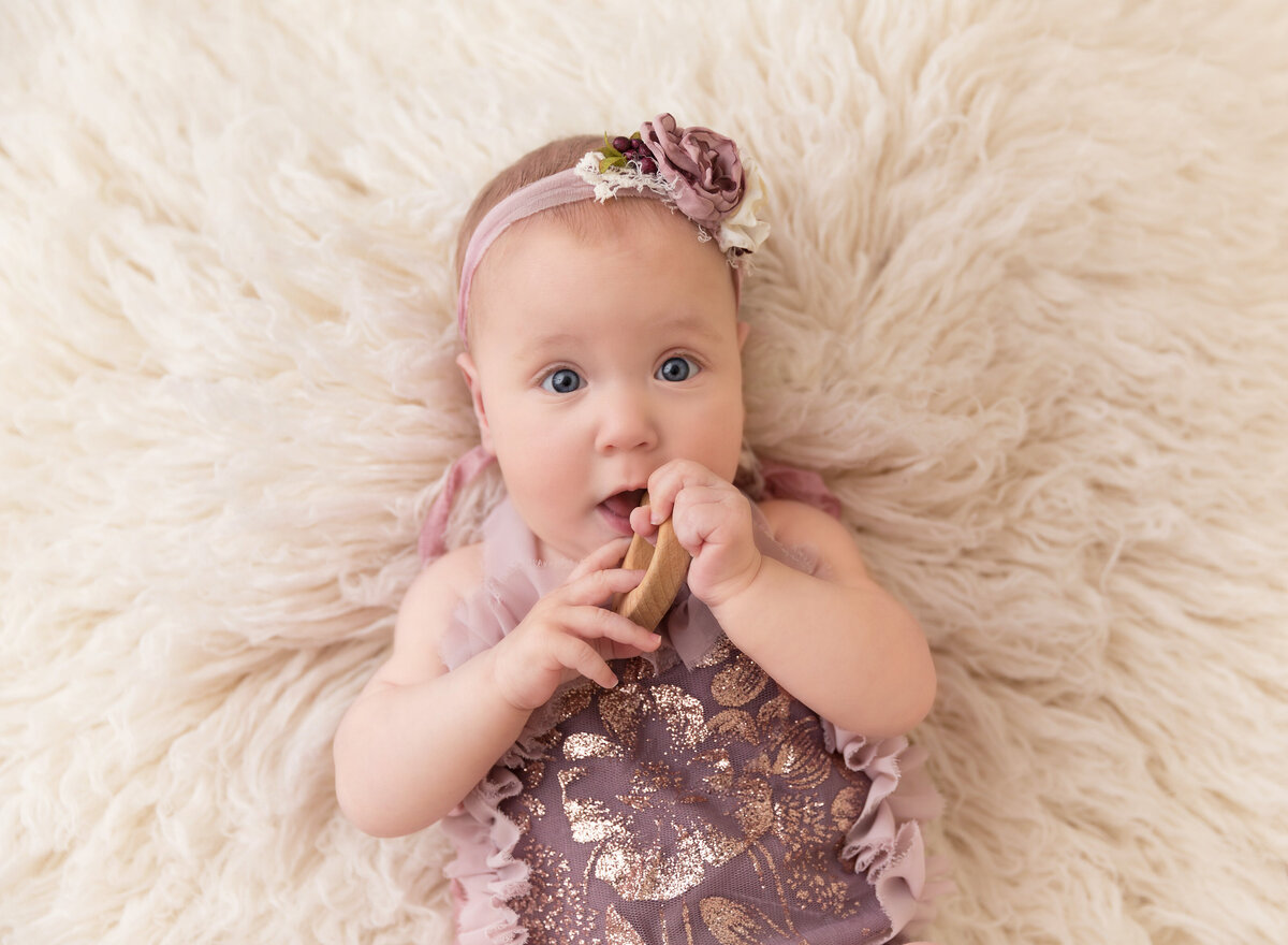 Baby girl is lying on her back for a 6-month baby milestone photoshoot. Aerial image. Baby is wearing a purple romper and holding a wooden toy.