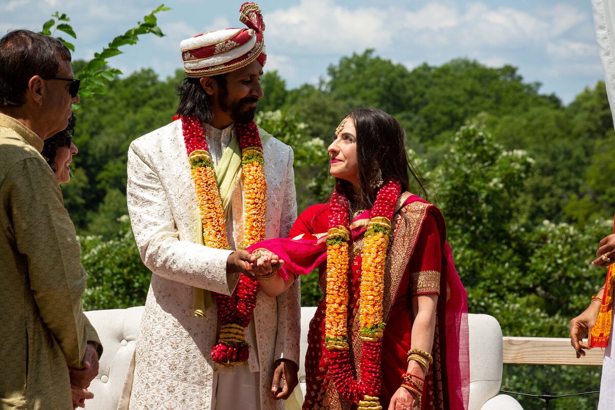 A bride and groom in traditional Indian attire holding hands at their wedding ceremony outdoors in Iowa, surrounded by guests.