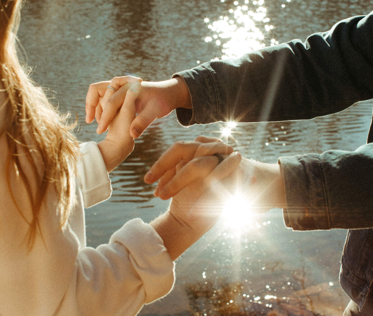 A couple is intertwining their fingers together and holding hands while they stand in front of a lake with the sunlight reflecting off the water.