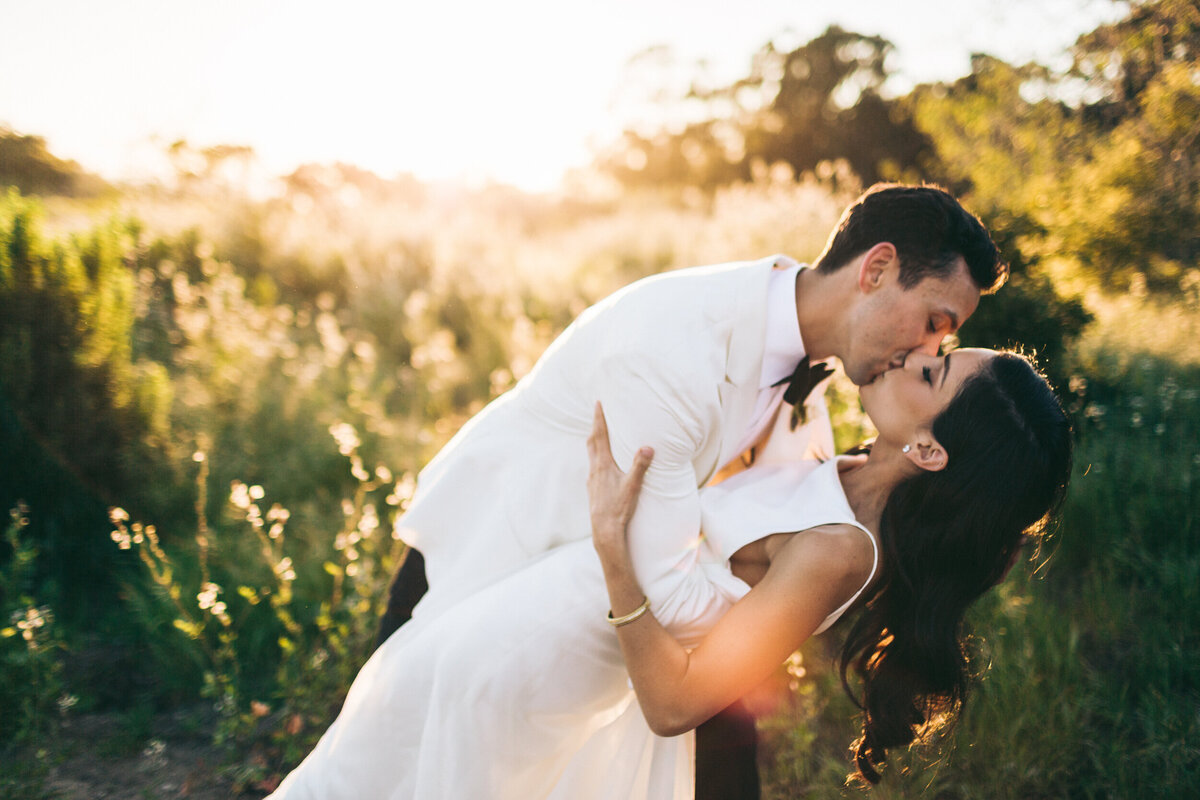 A colorful photograph of Sarah and Erik on their wedding day at Dos Pueblos Orchid Farm in Santa Barbara, California. The groom is dipping his bride while they embrace and kiss. The portrait is taken at sunset with the sun glowing behind them, lighting up the natural green grasses in a warm glow. He is in a tuxedo with white jacket and black pants and she is in a white satin wedding dress with a high neckline and narrow straps. Her dark, wavy hair is falling behind her and glowing from the sunset. Wedding photography by Stacie McChesney/Vitae Weddings.