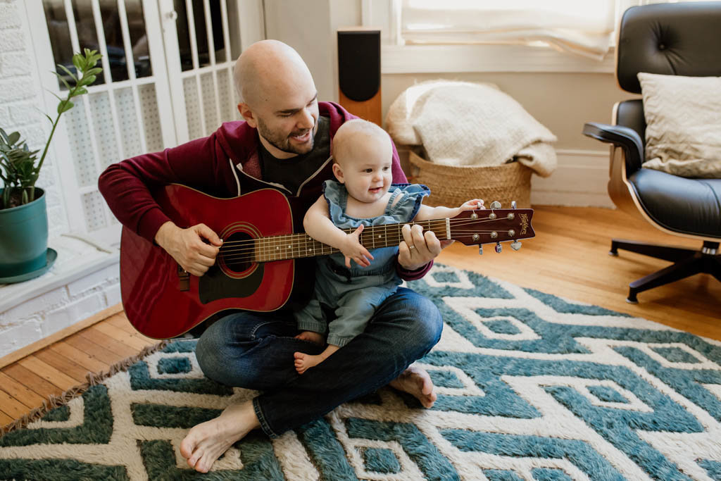 Lifestyle in home family photography session of dad playing guitar with daughter on lap