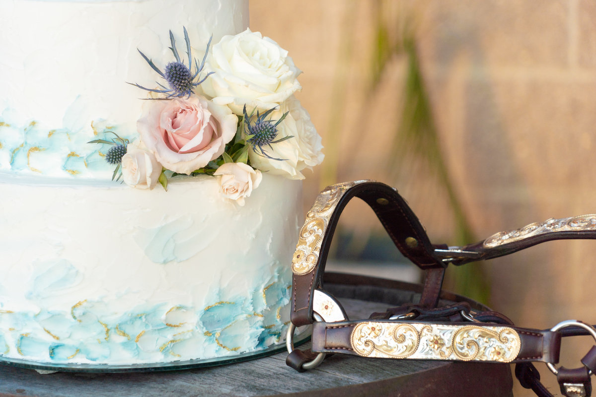 White wedding cake with light pink and white roses and teal markings next to a leather and gold bridle
