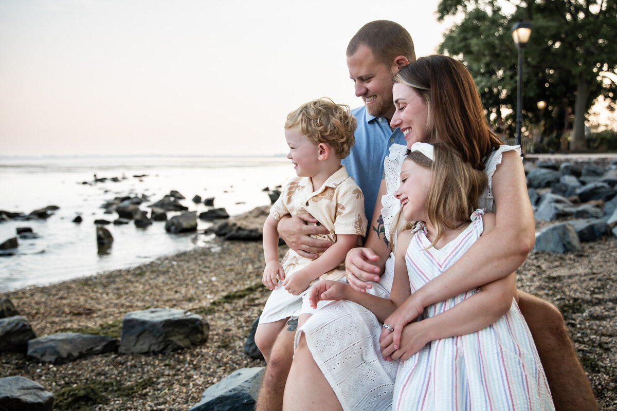 Family smiles while cuddling on beach near the Havre de Grace Promenade during their lifestyle family session with photographer