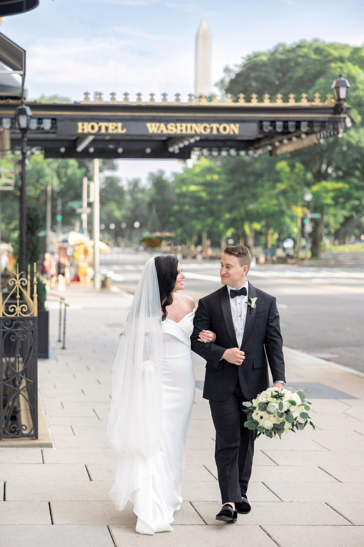 A bride in a white dress and groom in a black tuxedo walk arm-in-arm on a paved walkway. The bride holds a bouquet of white flowers. They are in front of a building marked Hotel Washington, with a monument visible in the background.