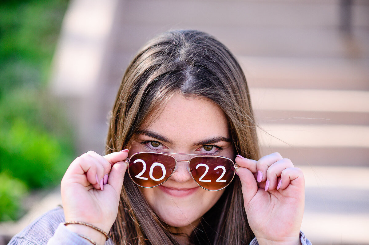 A Littleton senior photographer captures a girl with brown hair holding aviator sunglasses that say 2022 on them over her face and looks at the camera over the glasses.