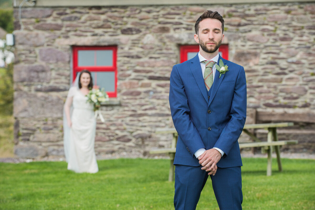 Groom wearing a blue wedding suit and green, tweed tie waiting for a first look of his bride who is wearing a vintage, beaded wedding dress holding a white and green bouquet outside an old stone cottage at Westcove House, Kerry