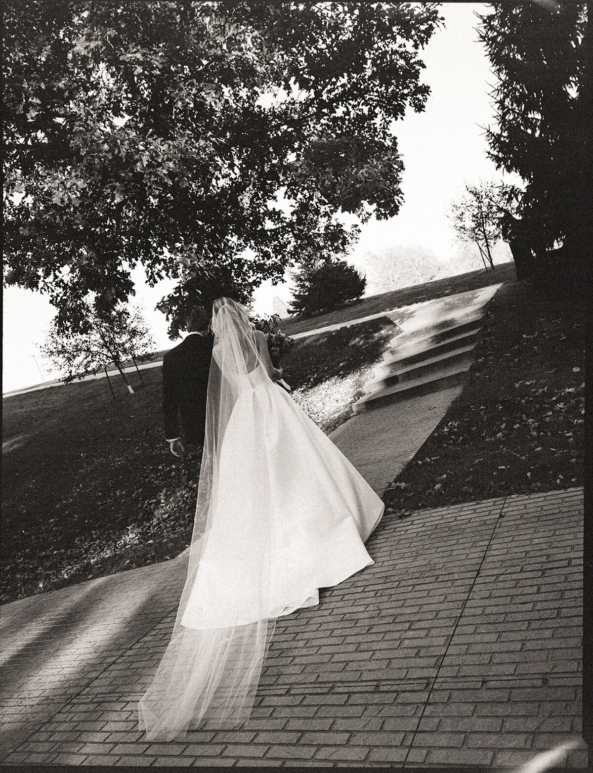 A bride in a flowing gown and veil walks hand in hand with a suited groom along a paved path, surrounded by trees and soft sunlight during an Iowa wedding.