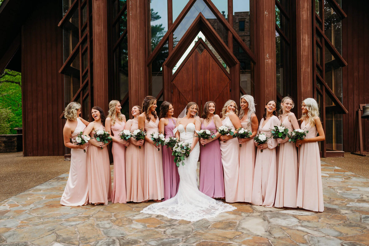 Photo of a bride laughing with her bridesmaids who are wearing pink in front of a wooden wedding chapel