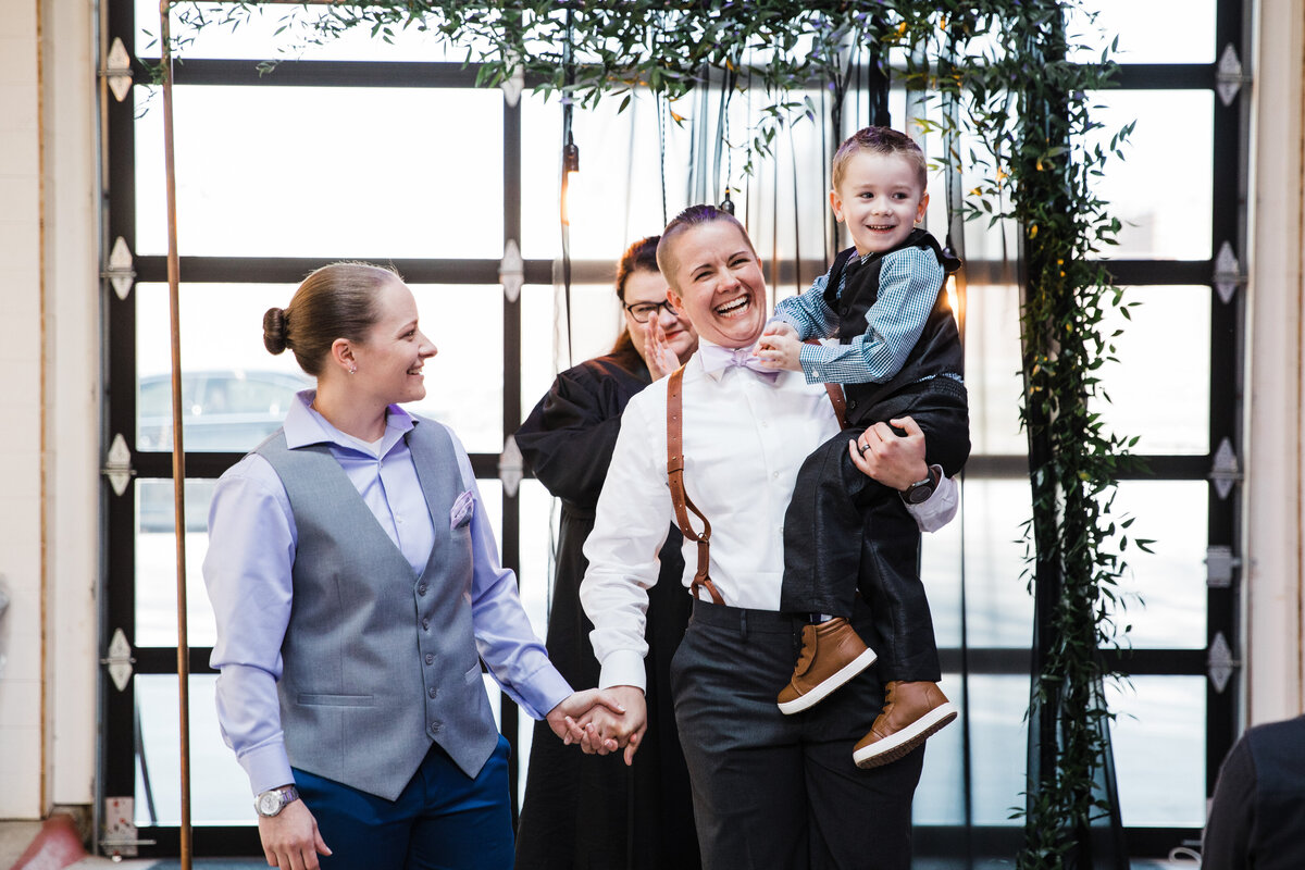 A candid shot of two brides and their child holding hands and getting ready to celebrate and process down the aisle after their wedding ceremony in Dallas, Texas. The bride on the left is wearing a lavender dress shirt, grey vest, and blue dress pants. The bride on the right is wearing a white dress shirt, bowtie, suspenders, and dark dress pants. Their child is being held by the bride on the right, and he's wearing formal attire as well. Their officiant can be seen in the background, and they're all in front of a large window.