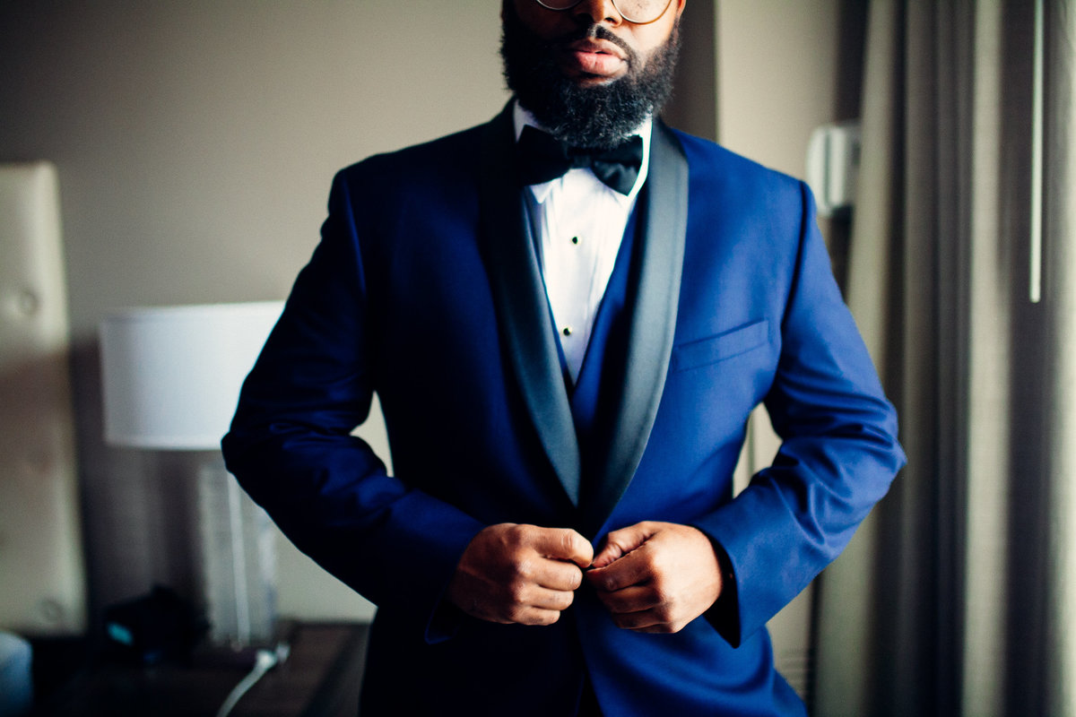 Groom gets ready to see his bride, buttoning up his blue suit jacket after making sure his bow tie is perfect.