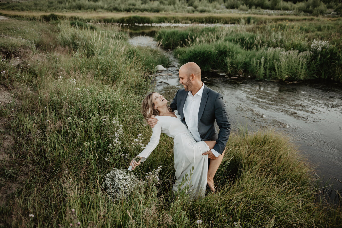 jackson hole wedding with groom dipping his bride back as she laughs  and throws back her head as the groom holds his brides leg, they are standing in a tall grass field in Jackson Hole photographed by jackson wyoming photographer