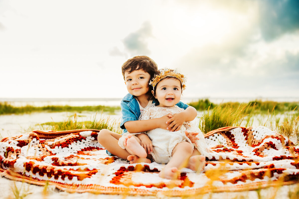 2 young kids sitting on a vintage blanket smiling at the camera