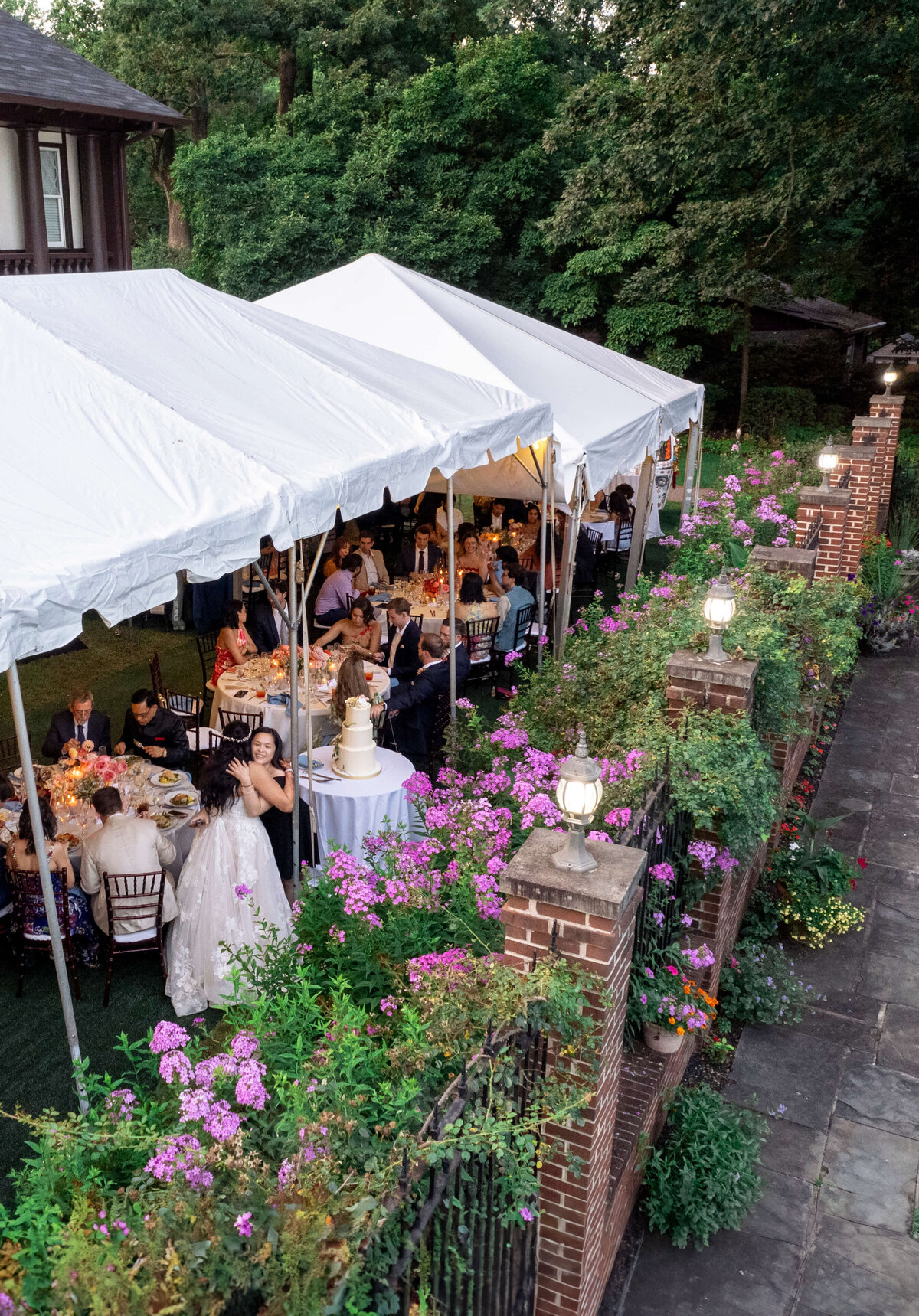 A wedding reception under a large white tent, surrounded by vibrant flowers and greenery. Guests are seated at round tables, and a couple is near a three-tiered wedding cake. Warm evening lights illuminate the scene.