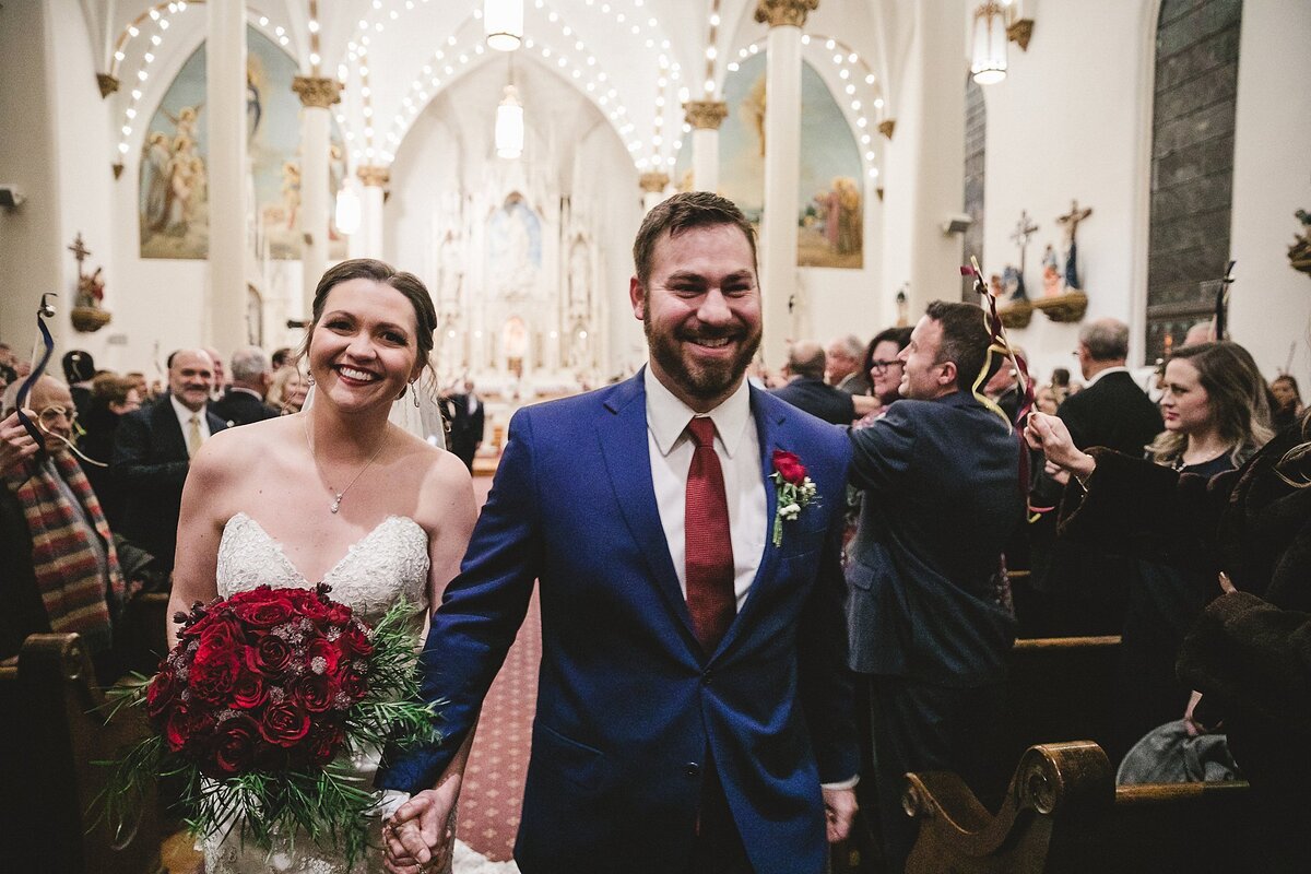 Bride wearing a strapless wedding dress, holding a round bouquet of red heart roses walks hand in hand with the groom wearing a blue suit with a red tie as the leave their wedding at a Nashville church.