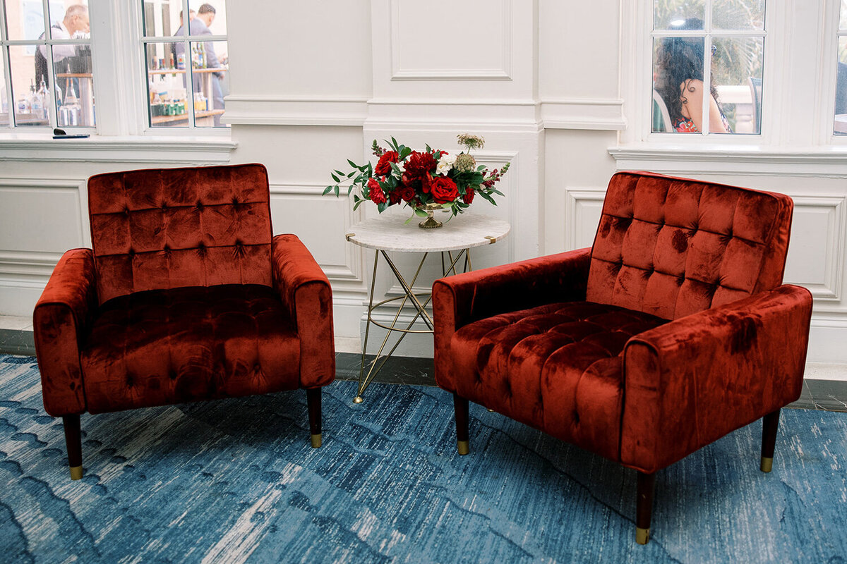 Chic lounge area at a December wedding reception at Don Cesar Hotel, St. Pete Beach, FL, featuring red velvet chairs, a white marble table, and red florals in a gold vase.